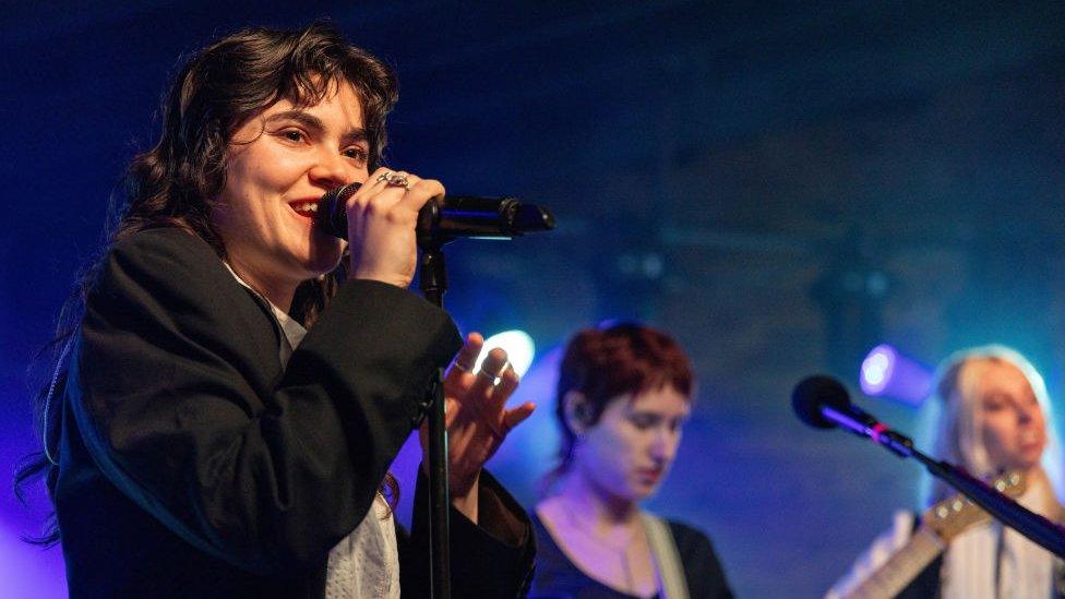 The Last Dinner Party performing in Austin, Texas in March 2024. Singer Abigail Morris is pictured in the foreground with Lizzie Mayland and Georgia Davies behind her. Abigail is a white woman in her 20s with long curly brown hair worn loose. She has brown eyes and wears an oversized black blazer over a white shirt. Lizzie is a white woman with short red hair and Georgia is a white woman with long blonde hair.