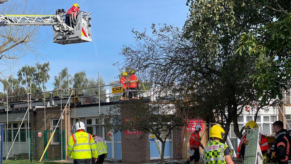 A firefighter in a crane lift above a low-lying school building with two firefighters on the roof and six others milling around below after a fire