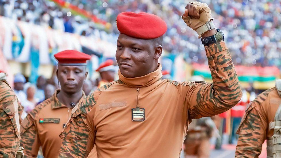 Ibrahim Traoré in beige military fatigues and red beret, surrounded by several soldiers in similar dress, pumps his fist in a stadium of his supporters.