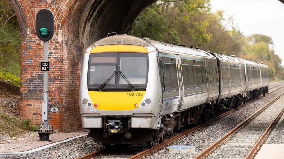 A chiltern class 168 travelling on the newly built east west rail route. It is travelling under a red brick bridge.