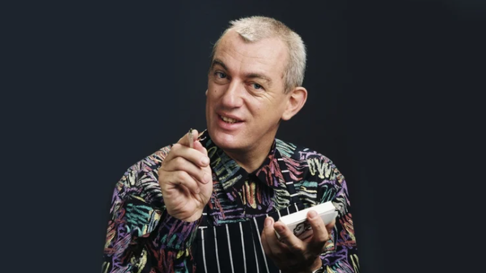 Shay Healy, holding a pen in his right hand and looking at the camera, wearing a black shirt with bright pastel coloured markings on it