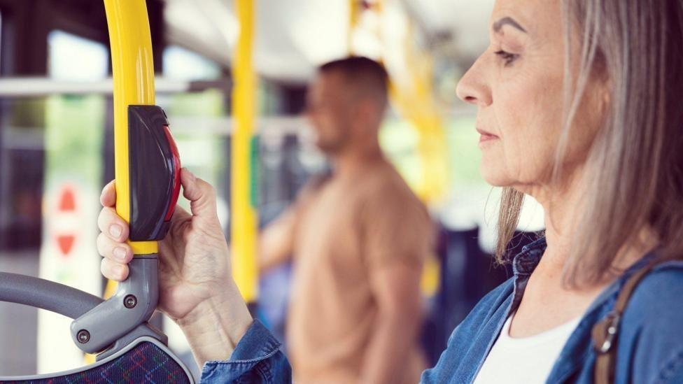 A woman presses the red stop button as she stands on a bus. A man can be seen standing in the background.