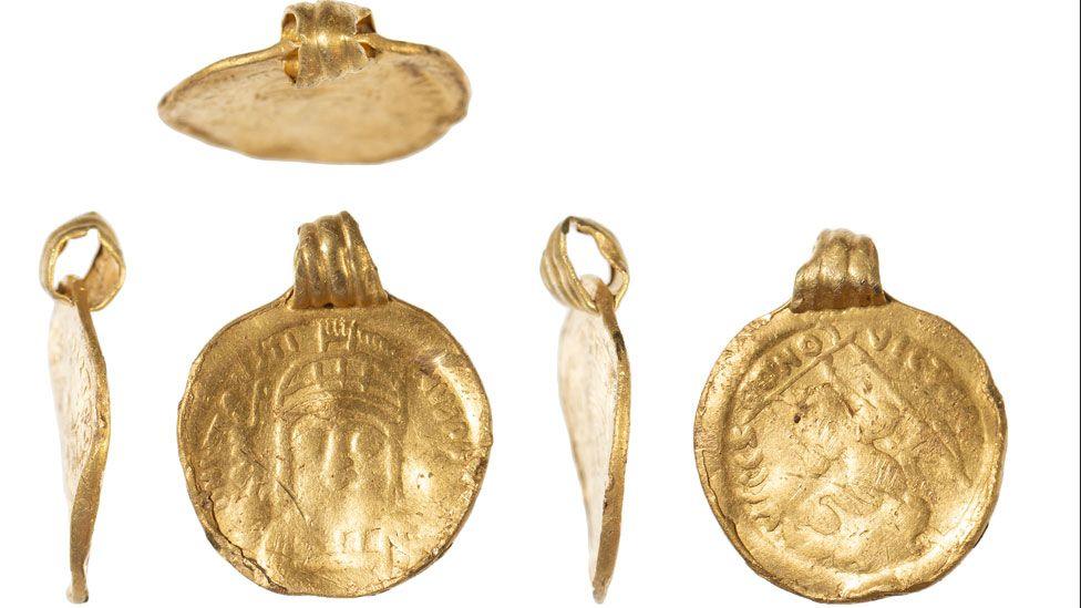 Five views of a 6th Century gold coin pendant. The first view above shows the pendant from above, looking at its hanging loop, made of five ribs of gold. Below are four views, the first showing it side on from the left, so light can be seen through the loop and showing the slight curve to the coin; the second shows the front of the coin, showing the face of Byzantine emperor Justin II, wearing a helmet with letters its circumference; the fourth shows a side on view of from the right, o light can be seen through the loop and showing the slight curve to the coin; the final view shows the reverse of the coin, showing Constantinopolis seated facing, looking right, holding sceptre and cross on globe and inscribed with the letters VICTORI-A AVGGG  S around its circumference.