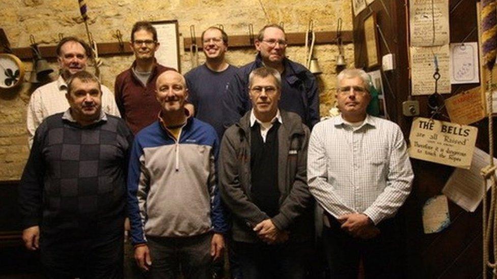 Eight man standing in two rows in a ringing chamber with bell ropes visible