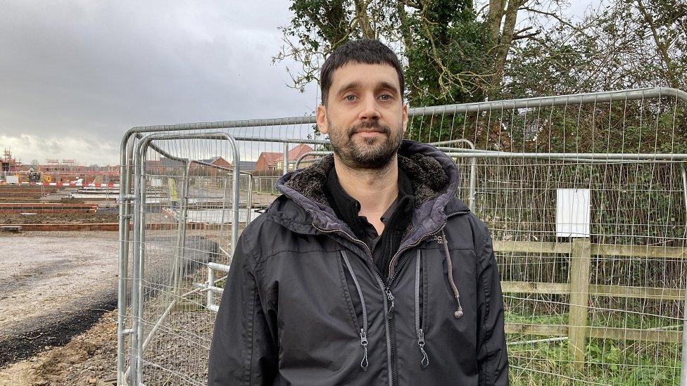 Sam Tibble looks at the camera in a winter coat in front of a construction site