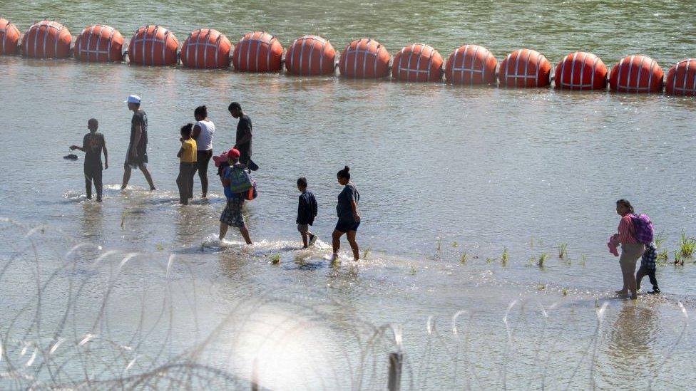 Migrants walk by a string of buoys placed on the water along the Rio Grande border with Mexico in Eagle Pass, Texas, on July 15, 2023, to prevent illegal immigration entry to the US.