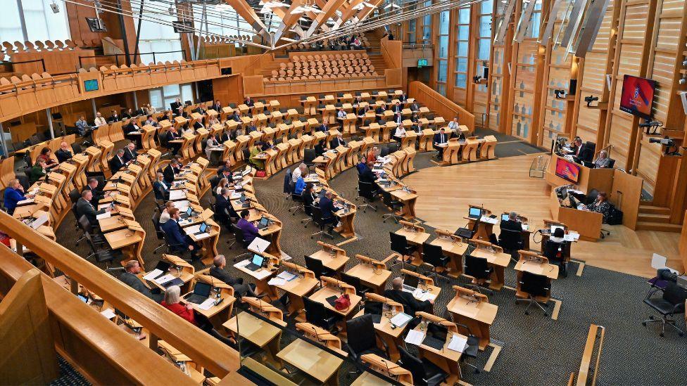 A general view of the Scottish Parliament chamber, with MSPs sitting at wooden desks in a semi-circle around a wooden speaker's podium.