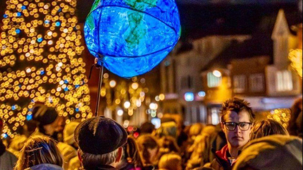 Blurred heads in a crowd with a lantern in the shape of the Earth held aloft and the gold and blue lights of a Christmas tree to left.