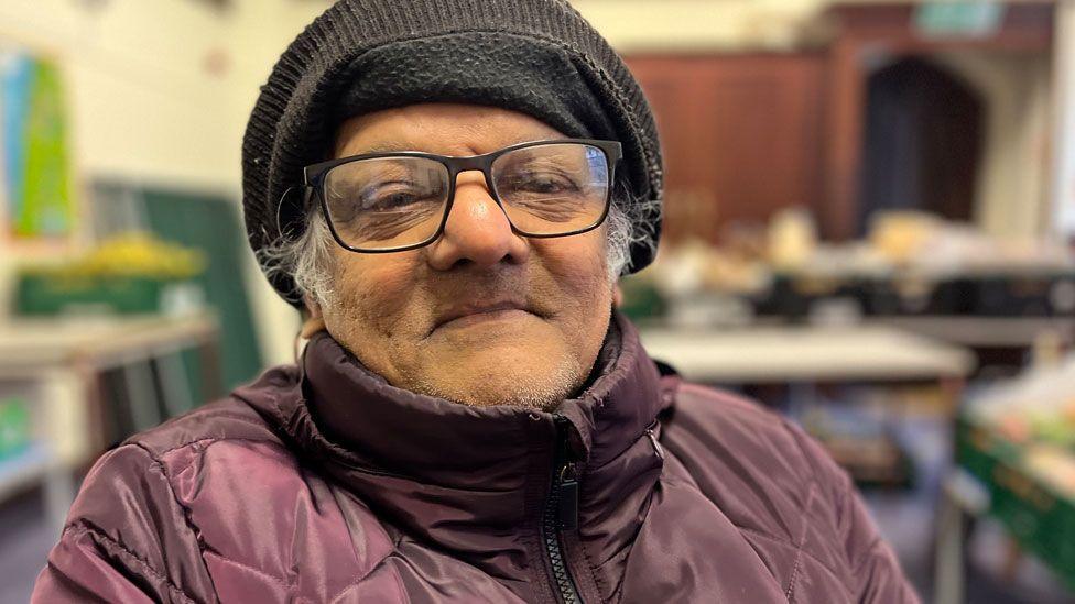 Stanley Coutinho who is sitting down in a community-hall style building. He has a black fleece hat on his head and white hair can be glimpsed. He is wearing black-framed glasses and and a purple padded jacket, zipped up to the neck.