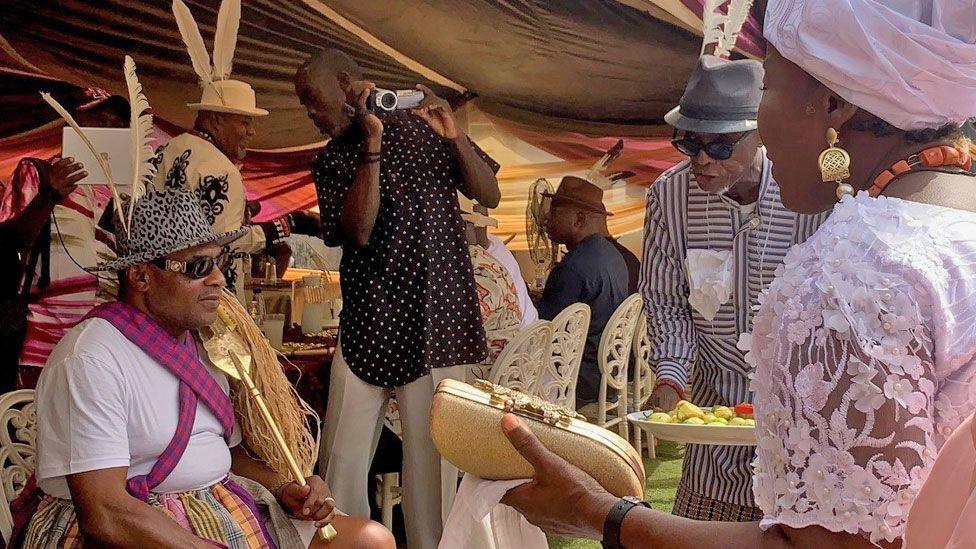 Pascal Okey Adizua is seated, wearing a leopard print hat with two feathers attached to it. He is being approached by a someone carrying  a plate of food and a woman who is attending his Ogbuagu ceremony