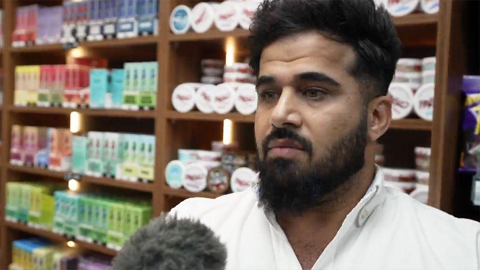 Close-up of Twana Jamal with dark hair and a full beard, wearing a white collared shirt, standing indoors in front of wooden shelves filled with neatly arranged, colourful product boxes and small round containers. A microphone with a fuzzy windscreen is positioned in the lower foreground, indicating an interview or recording. The background features warm lighting within the shelving units, highlighting the rows of packaged items.