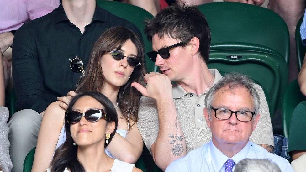 Daisy Edgar-Jones and Ben Seed sit side-by-side, and Seed whispers something to Edgar-Jones as she tilts her head in his direction. Both are wearing sunglasses.