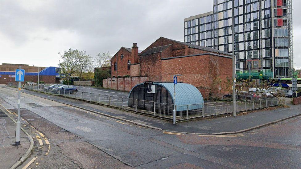 Street view image of the proposed plot. A car park by a junction with  red brick building in the background and a round shelter to the front