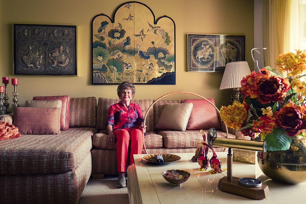 Photo shows Joan Anderson in a bright pink button down shirt and pink trousers holding a wooden hoola hoop. She smiling while sat on a striped sofa in  a yellow room with flowers and glass ornaments on a coffee table and paintings on the wall