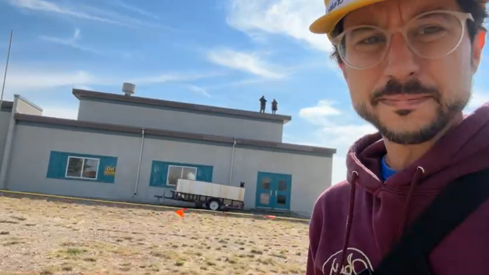 Thomas Fougere of Community TV reporting from outside the school while two followers of Ms Didulo, standing on the roof, look on