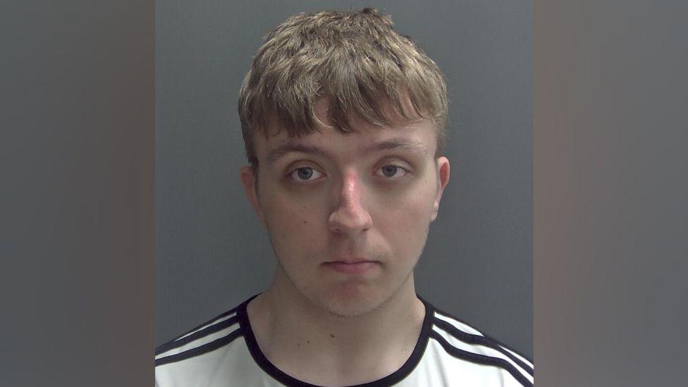 Samuel Gorton is seen looking at a camera in a police custody shot. He has short, light brown hair cropped above his ears, with a fringe. He is wearing a white T-shirt with black stripes around the neckline and shoulders