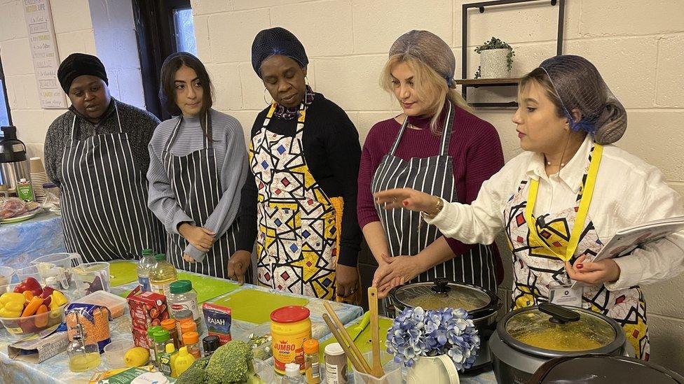 Two teachers take a cooking class with three participants. They are Standing behind a table filled with food.