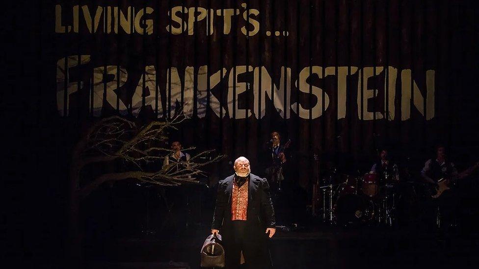 Howord Coggins stands in the middle of a stage as Victor Frankenstein, with the name of the play at the back of the dark coloured set and dressed in a three-piece suit with cravat.