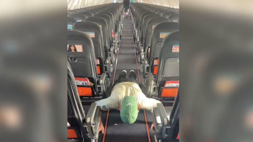 Alex Welford wearing a white top, black trousers and a green cap on backwards, doing a press up in the aisle of a plane