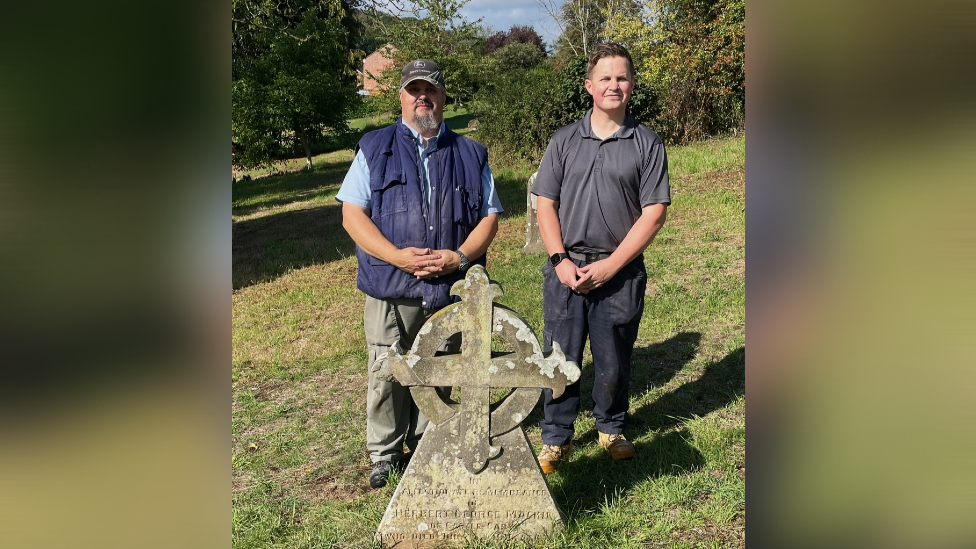 The picture shows Martin Adams and Phil Curnow standing behind the headstone for Herbert Mackie.  There are trees and grass in the background.