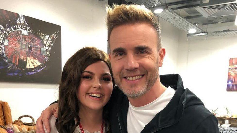 Molly Cuddihy is smiling in close up, with red beads round her neck and a white blouse. Gary Barlow in black jacket and white T-shirt is standing beside her with his right arm around her.