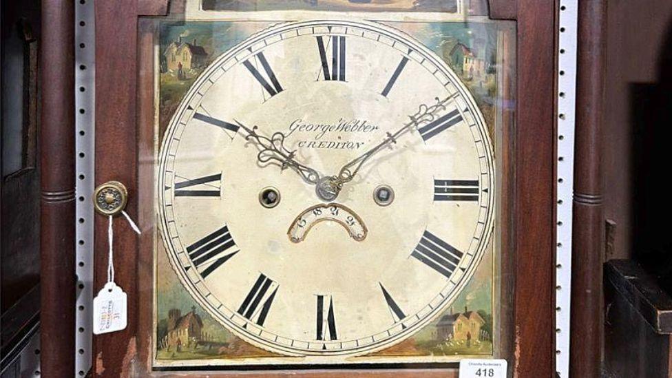 A brown wooden clock with a painted face that depicts houses and green grass and a boy above the face. The picture of the clock is on a light purple background.