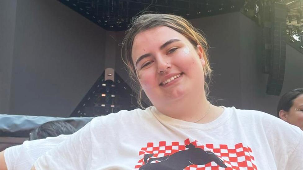 Harriet smiling looking at the camera with her head slightly tilted. She is wearing a white tshirt with red and black squared patterns. The background is of black speaker staging.