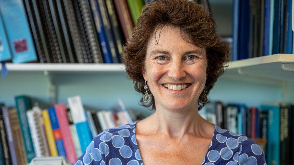 Prof Rebecca Fitzgerald sitting in front of two shelves packed with books and files. She has short dark curling hair, is wearing a blue top and is smiling 