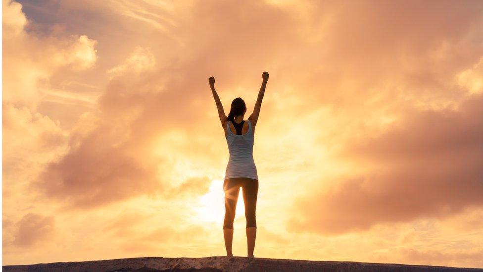 A woman in gym kit standing on a hill in the sun celebrating