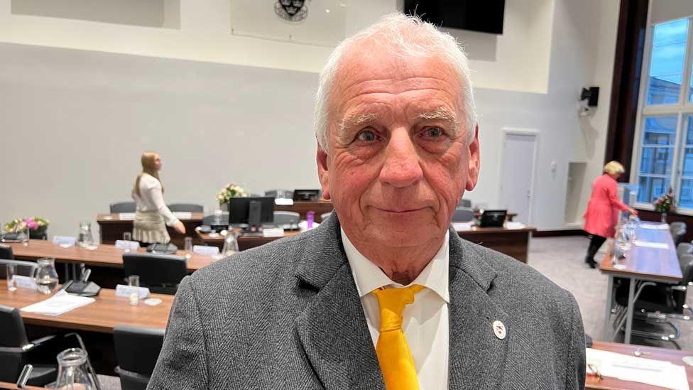 Barry Aspinell inside a council meeting chamber, which has chairs and tables behind him. He has short grey hair and is smiling at the camera. He is wearing a grey suit with a white shirt and yellow tie.