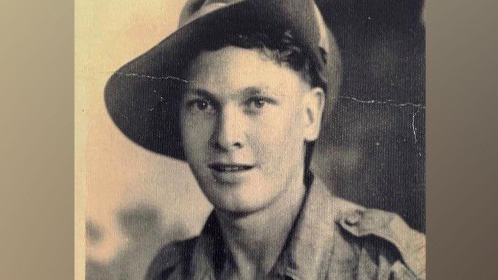 A sepia-toned black and white head and shoulders shot of Charlie Richards wearing Army uniform. He has a broad brimmed hat on and is wearing an open necked shirt. He has short hair and is starting to smile. 