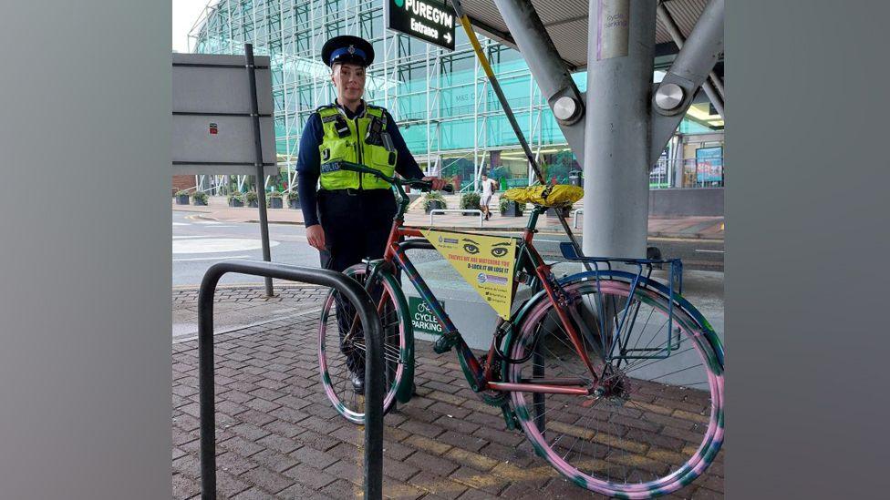 A female police officer stands next to a bike that has brightly coloured wheels with pink and green flecks. There is a yellow sign on the bike with warning messages to thieves and cyclists and the saddle is bright yellow. The bike is chained to a cycle rack outside a shopping centre, with large glass panels that are coloured slightly turquoise. The officer wears a high visibility vest over a blue uniform and a black and blue police hat.