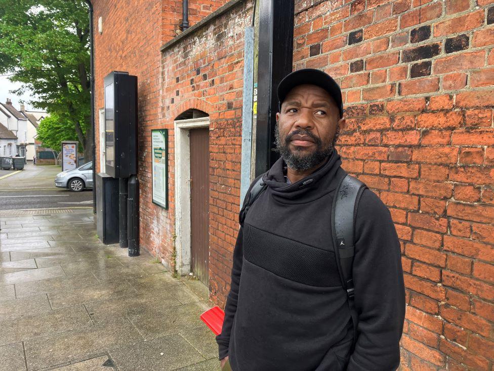 Ignatius is standing on a pavement dressed all in black. He is standing in front of a brick wall.