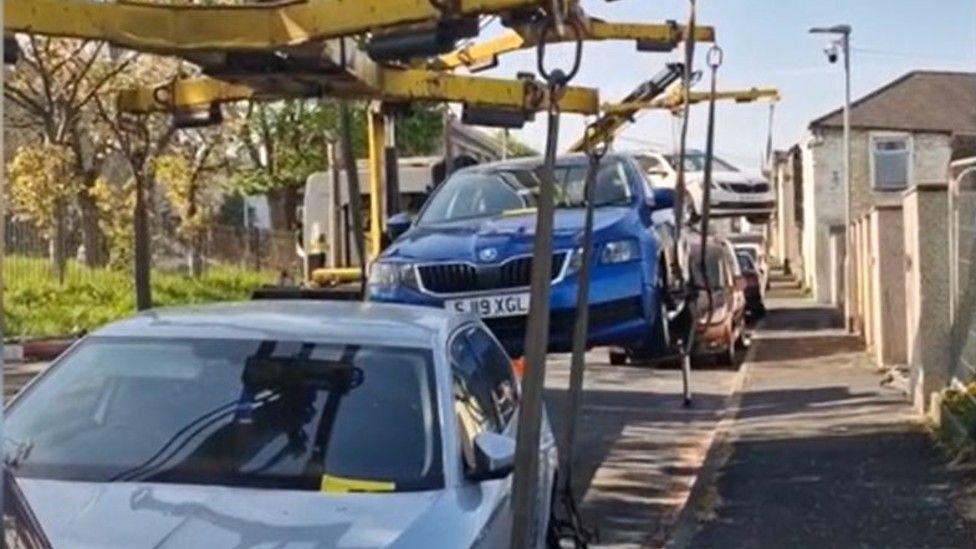 A line of cars being hauled away by a large grabbing machine.