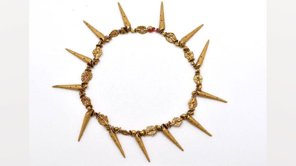 A 19th Century necklace from Asante, Ghana. It is intricately worked in rich yellow gold with flat and round gold beads strung along in a circle, and 13 long gold spikes shaped like spiral shells spacing out the beads every seventh bead