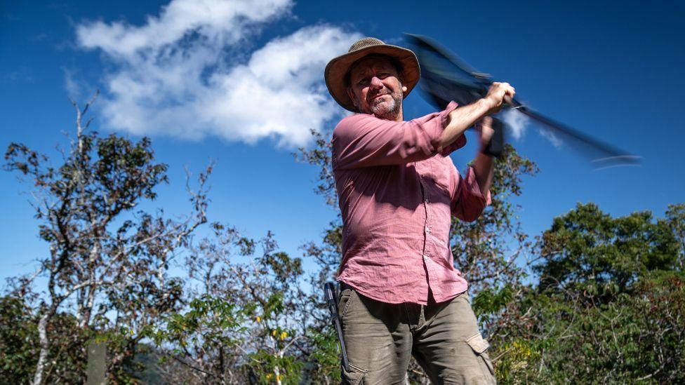 man in widebrimmed hat swirling a butterfly net with forest and blue sky in background