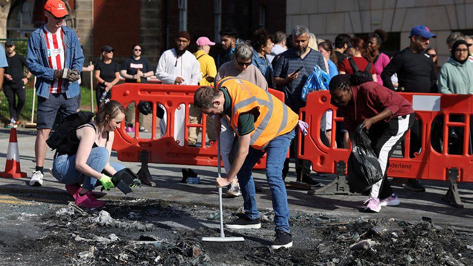 People help sweep up debris from a burnt vehicle after a night of violent anti-immigrant demonstrations in Sunderland