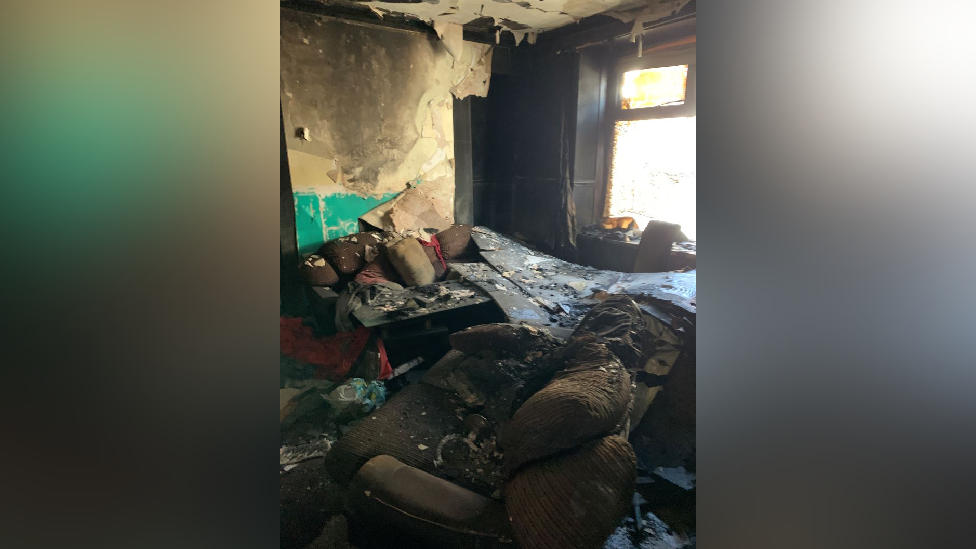 The inside of the same terraced house in another room, with new debris including a burnt and dirt covered sofa and peeling burnt wallpaper 