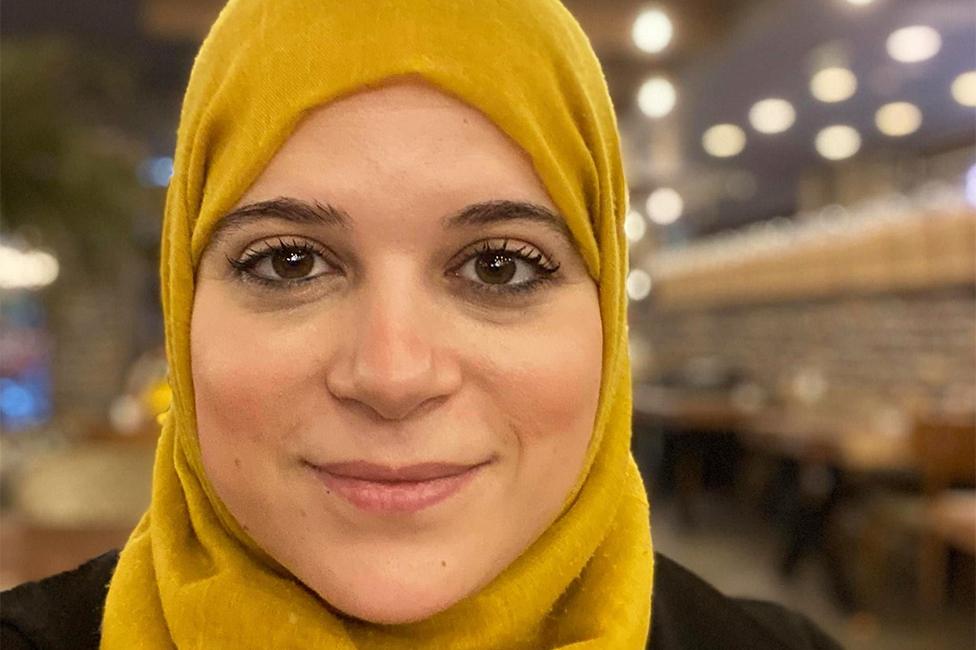 Amna in a selfie photo, smiling while wearing a yellow hijab - which is an Islamic head covering - while the background is blurred.