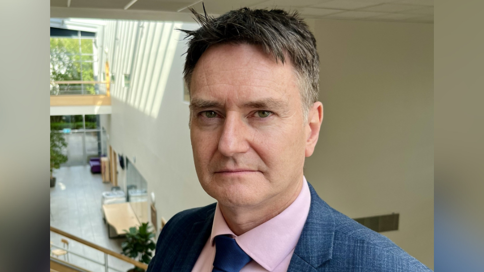 Head of organ transplantation at NHS Blood and Transplant, Anthony Clarkson. He is stood upstairs overlooking a building atrium. He is wearing a blue suit, pink shirt and blue tie. He is looking directly at the camera.