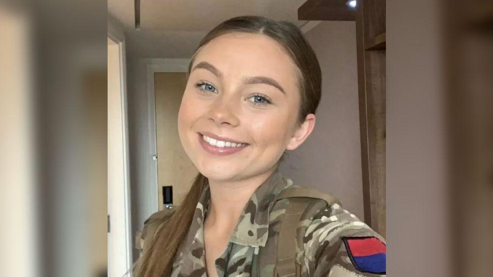 A selfie of Jaysley Beck wearing her military uniform, with her long brown hair slicked back into a low ponytail. She has blue eyes and is smiling at the camera.