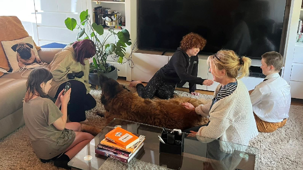 Four teeangers, an adult woman and a dog sat on a carpet. The kids are chatting to eachother and the woman pets the dog. There is a sofa , a TV and a bookshelf behind them and a glass coffee table in the forefront of the picture.