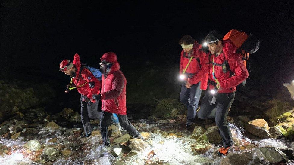 Four people in red coats walk through a stream in the dark