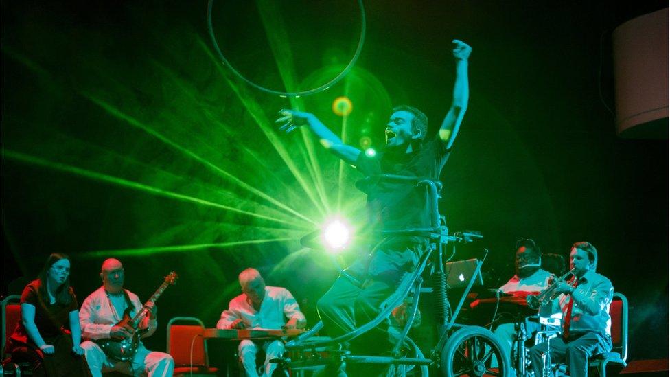 A disabled performer on stage with musicians performing in the background