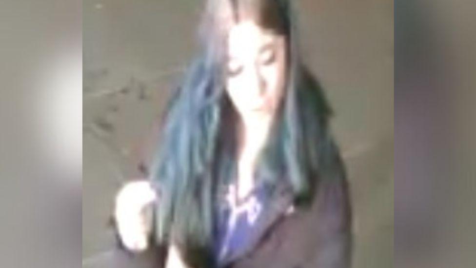 Another CCTV picture of the woman, who is looking down with her mouth open.  The picture is very over exposed, and she appears to be wearing a dark jacket with a blue top underneath. 