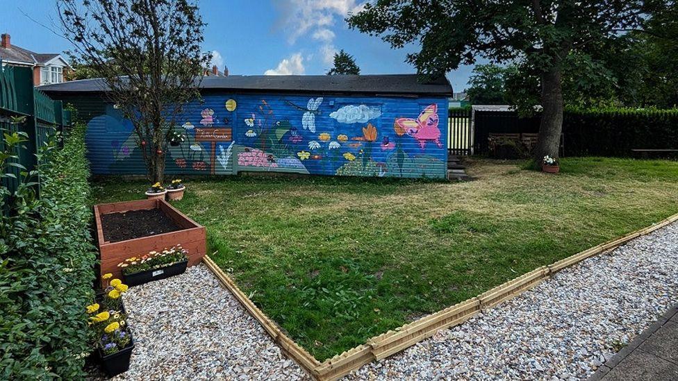 A large shed with a mural depicting flowers and insects on a square lawn with a pebble path and borders of flowers and trees