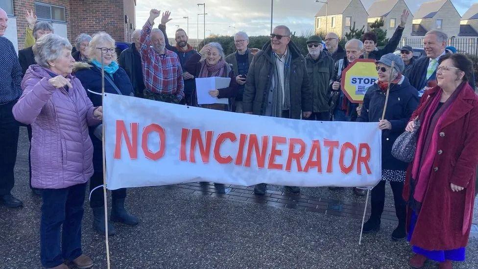 A group of people, some with their fists in the air, stand around a large banner reading 'no incinerator'