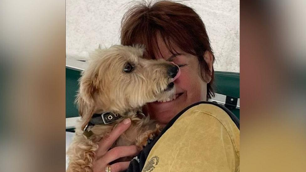 Isobel Heely wearing a yellow top with short brown hair, smiling and holding a dog in front of her face