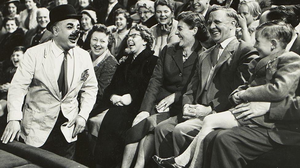 Black and white image of Charlie Cairoli ringside by a crowd of laughing people looking towards a laughing child. He is wearing a bowler hat, white jacket, shirt and tie and has a big clown nose, large eyebrows, a moustache and dark lips