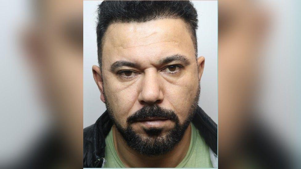 Mugshot of Mukhlis Jamal Hamadamin. He has black hair and a trimmed beard. He is photographed wearing a green t-shirt and black jacket.